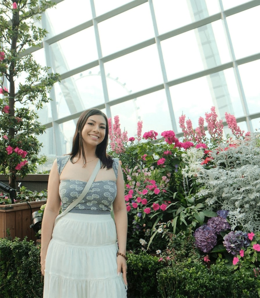 Lac Nhi smiling in a lush conservatory among flowers and greenery, with the Singapore Flyer visible through a bright glass grid.